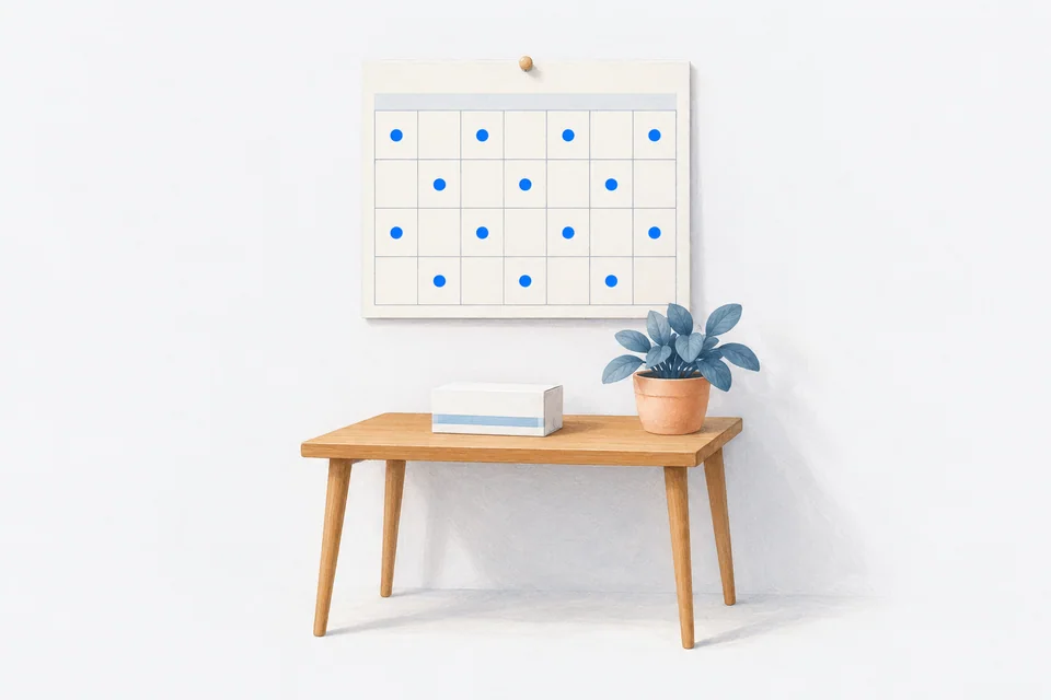 A blank wall calendar with alternating blue dots hangs above a small wooden desk with a medicine box and a potted plant.